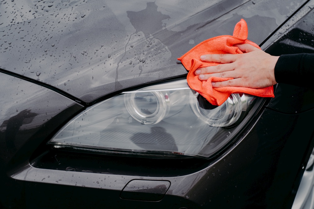 Mans hand with rag wiping cars headlamp. Wet black automobile cleanes at car wash. Auto cleaning and service concept