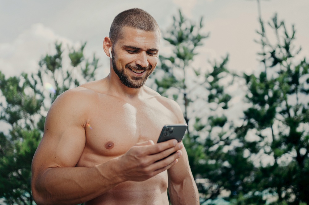 Cheerful motivated shirtless sportsman concentrated in smartphone, sends text messages, uses wireless earphones for listening music, has strong muscles, stands outdoor. Sporty lifestyle concept