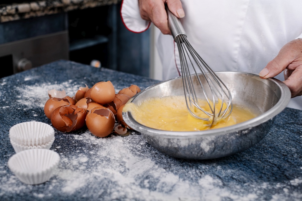 Close-up of a baker mixing eggs with a hand blender in a steel bowl.