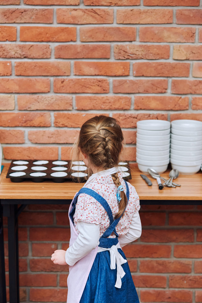 Little girl looking at cups for cakes. Kid taking part in baking workshop. Baking classes for children,  aspiring little chefs. Learning to cook. Combining and stirring prepared ingredients. Real people, authentic situations