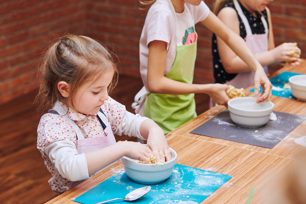 Little girls kneading the dough for baking the cake. Kids taking part in baking workshop. Baking classes for children,  aspiring little chefs. Learning to cook. Combining and stirring prepared ingredients. Real people, authentic situations