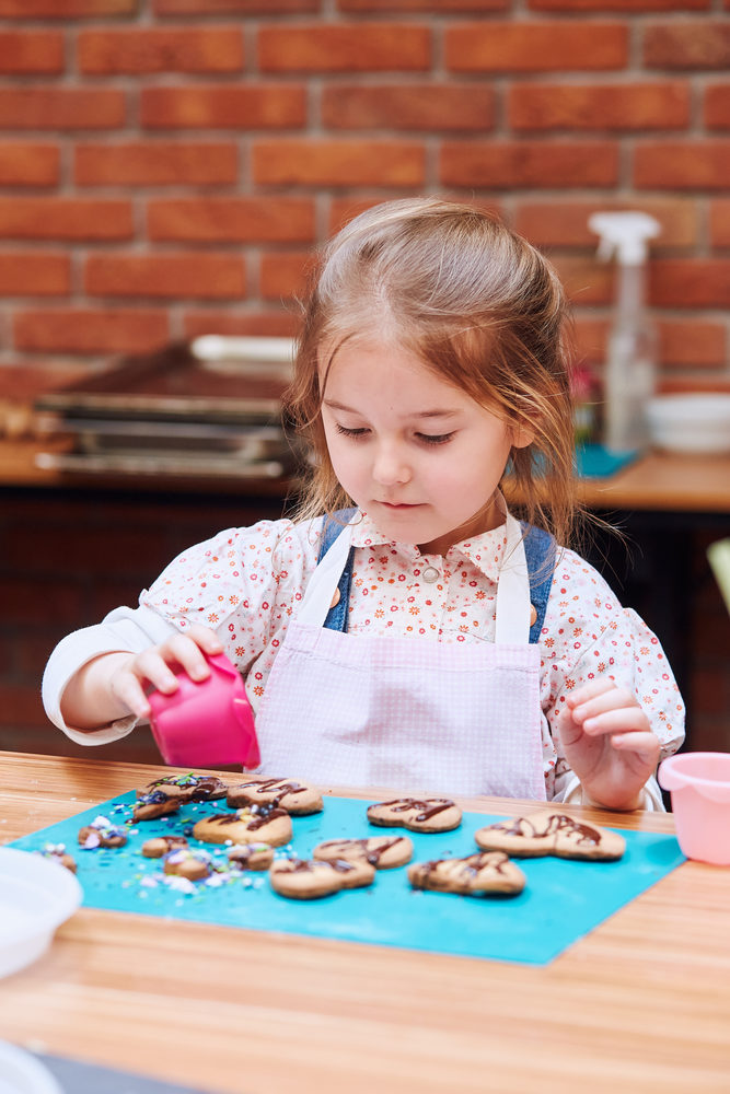Little girl decorating her baked cookies with colorful sprinkle and icing sugar. Kid taking part in baking workshop. Baking classes for children,  aspiring little chefs. Learning to cook. Combining and stirring prepared ingredients. Real people, authentic situations