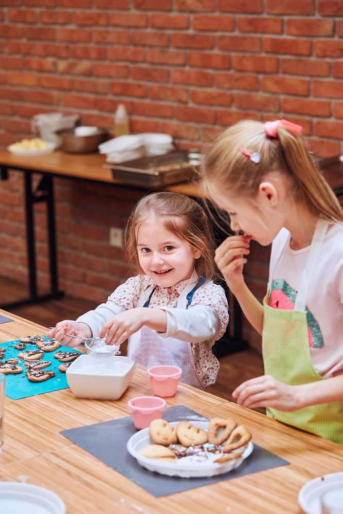 Little girls decorating baked cookies with colorful sprinkle and icing sugar. Kids taking part in baking workshop. Baking classes for children, aspiring little chefs. Learning to cook. Combining and stirring prepared ingredients. Real people, authentic situations