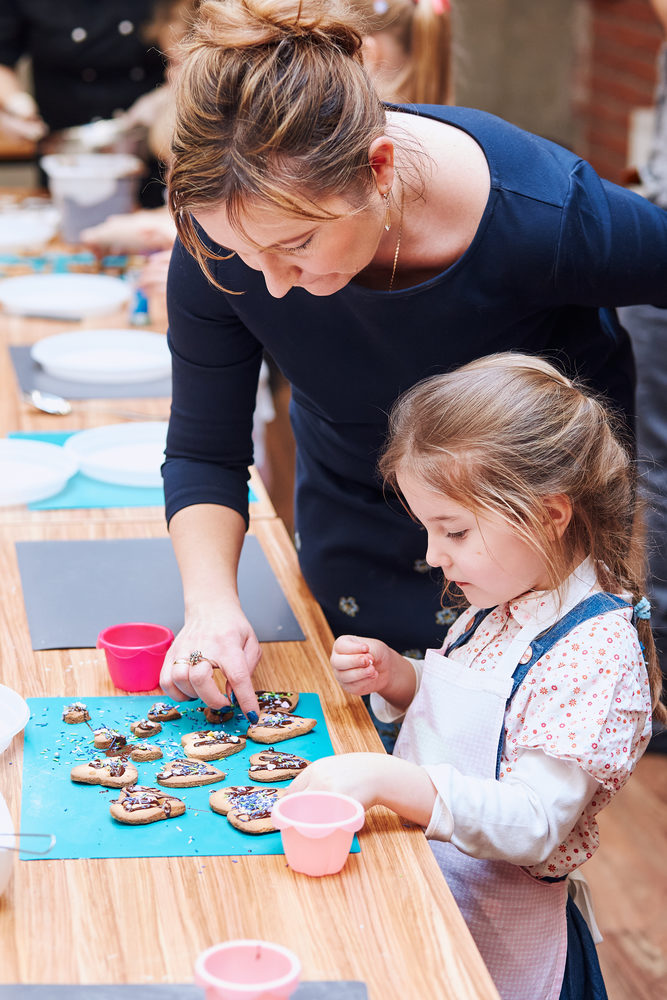 Little girl with her mom&rsquo;s help decorating baked cookies with colorful sprinkle and icing sugar. Kid taking part in baking workshop. Baking classes for children, aspiring little chefs. Learning to cook. Combining and stirring prepared ingredients. Real people, authentic situations