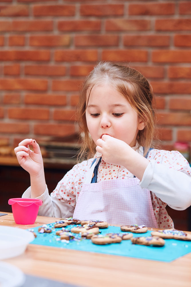Little girl tasting cookie baked oneself. Decorating her baked cookies with colorful sprinkle and icing sugar. Kid taking part in baking workshop. Baking classes for children,  aspiring little chefs. Learning to cook. Combining and stirring prepared ingredients. Real people, authentic situations
