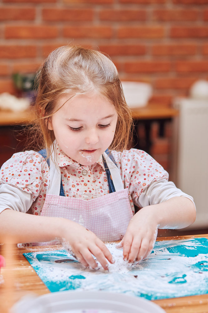 Little girl eating the icing sugar left after baking cookies. Kid taking part in baking workshop. Baking classes for children, aspiring little chefs. Learning to cook. Combining and stirring prepared ingredients. Real people, authentic situations