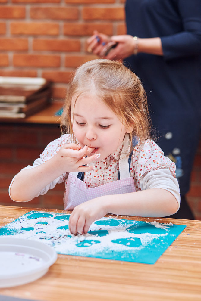 Little girl eating the icing sugar left after baking cookies. Kid taking part in baking workshop. Baking classes for children, aspiring little chefs. Learning to cook. Combining and stirring prepared ingredients. Real people, authentic situations