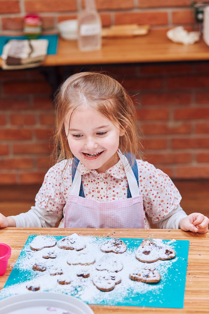 Little girl happy because of her baked cookies. Decorating cookies with colorful sprinkle and icing sugar. Kid taking part in baking workshop. Baking classes for children, aspiring little chefs. Learning to cook. Combining and stirring prepared ingredients. Real people, authentic situations