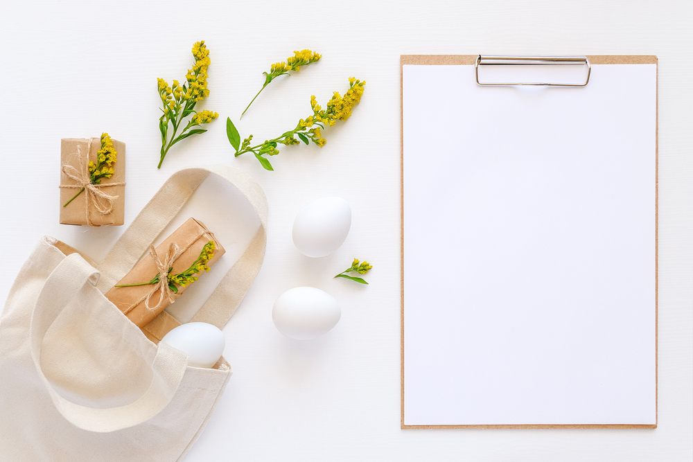 Happy easter concept. White eggs, yellow flowers, craft gifts fly out textile bag and clipboard with paper on white background Mockup Flat lay Top view.. Happy easter concept. White eggs, yellow flowers, craft gifts fly out textile bag and clipboard with paper on white background Mockup Flat lay Top view
