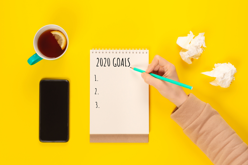 2020 goals banner. Notebook, pencil and cup of tea on a yellow background near crumpled sheet of paper and mobile phone. Copy space, Flat lay. Women hand write to do list for 2020 year. 2020 goals banner. Notebook, pencil and cup of tea on a yellow background near crumpled sheet of paper and mobile phone.