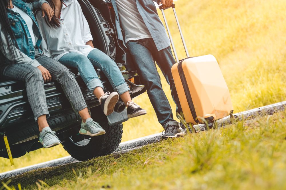Closeup lower body of group of friends relaxing on SUV car trunk with trolly luggage along road trip with autumn mountain hill background. Freedom  road way. People lifestyle transportation travel