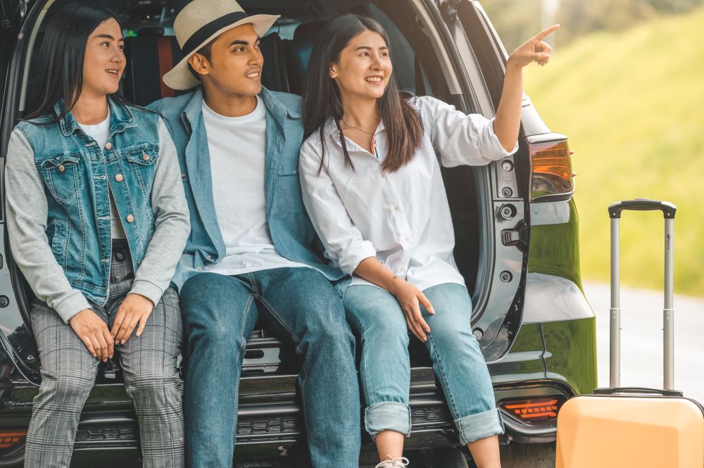 Asian man and women relaxing in back of car trunk during travel in summer. Transportation and people lifestyles concept. Group of happy friendship sit and looking nature view. Relaxation in vacation