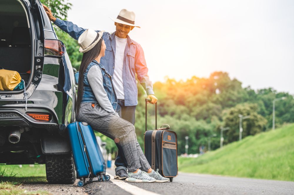 Closeup lower body of Asian couple relaxing on SUV car trunk with yellow trolly luggage along road trip with mountain hill background. Freedom road life. People lifestyle and transportation travel