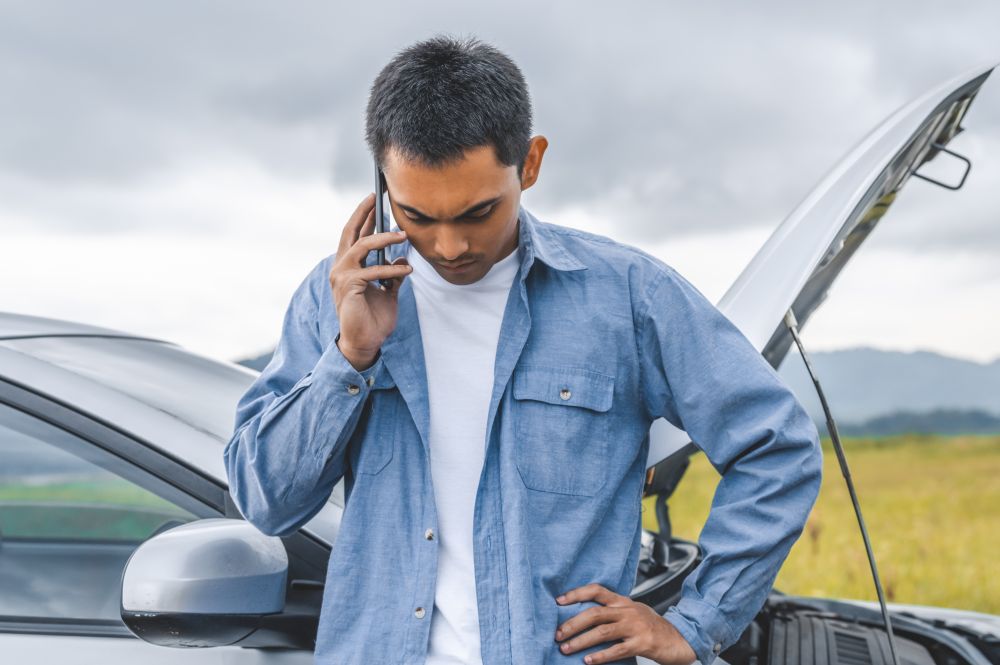 Asian man calling car maintenance service after having accident during travel long weekend between road trip. Car broken in mountain meadow background. People transportation. Need help insurance claim