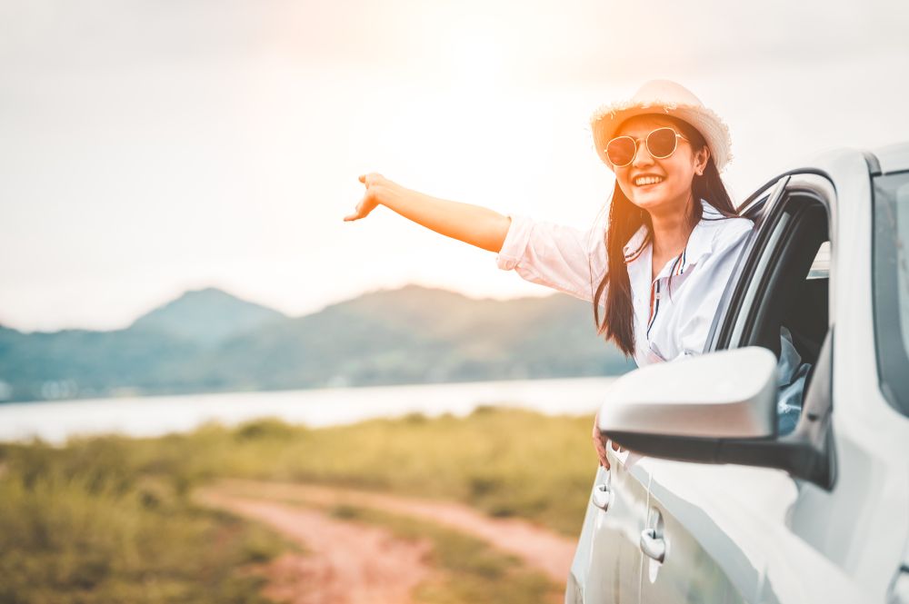 Happy woman waving hand outside open window car with meadow and mountain lake background. People lifestyle relaxing as traveler on road trip in holiday vacation. Transportation and travel concept