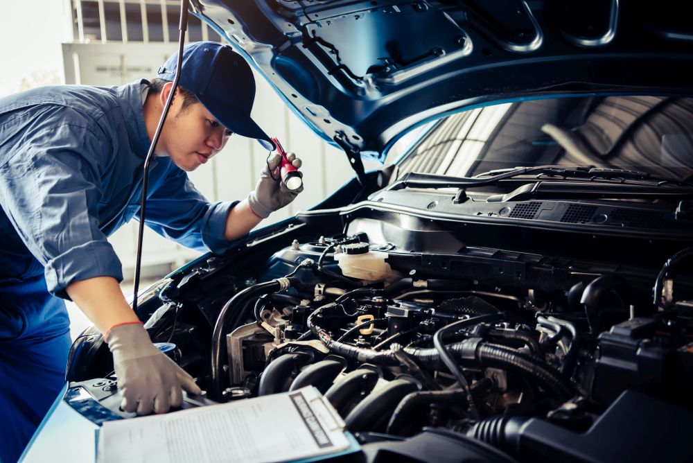 Car mechanic technician holding flashlight checking engine with checklist clipboard to maintenance vehicle by customer claim order in auto repair shop garage repair service. People occupation business