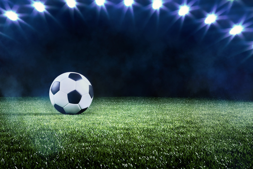 Football or soccer background with a row of spotlights illuminating a ball on the green turf in a stadium in a sports championship or World Cup event in a panorama banner
