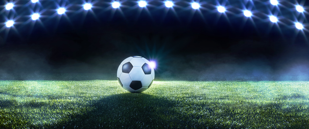 Football or soccer background with a row of spotlights illuminating a ball on the green turf in a stadium in a sports championship or World Cup event in a panorama banner