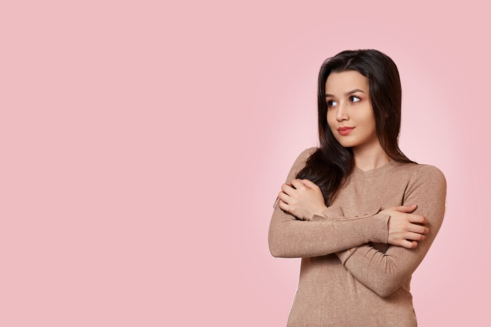 portrait of a young beautiful Asian brunette girl in a light brown jacket stands with her arms crossed on her chest . on a pink isolated background with copyspace.