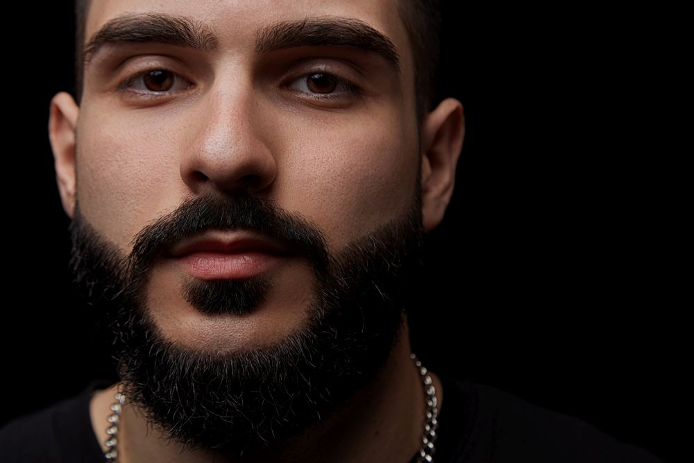 a close-up dramatic portrait of a young serious guy, musician, singer,rapper with a beard and arms crossed on his chest on a black isolated background.