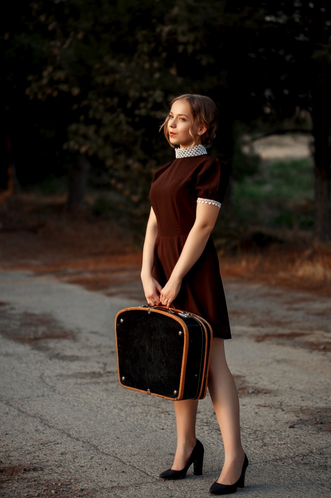 a young beautiful girl in a brown dress in a retro style stands with a black suitcase in her hands on an abandoned road.