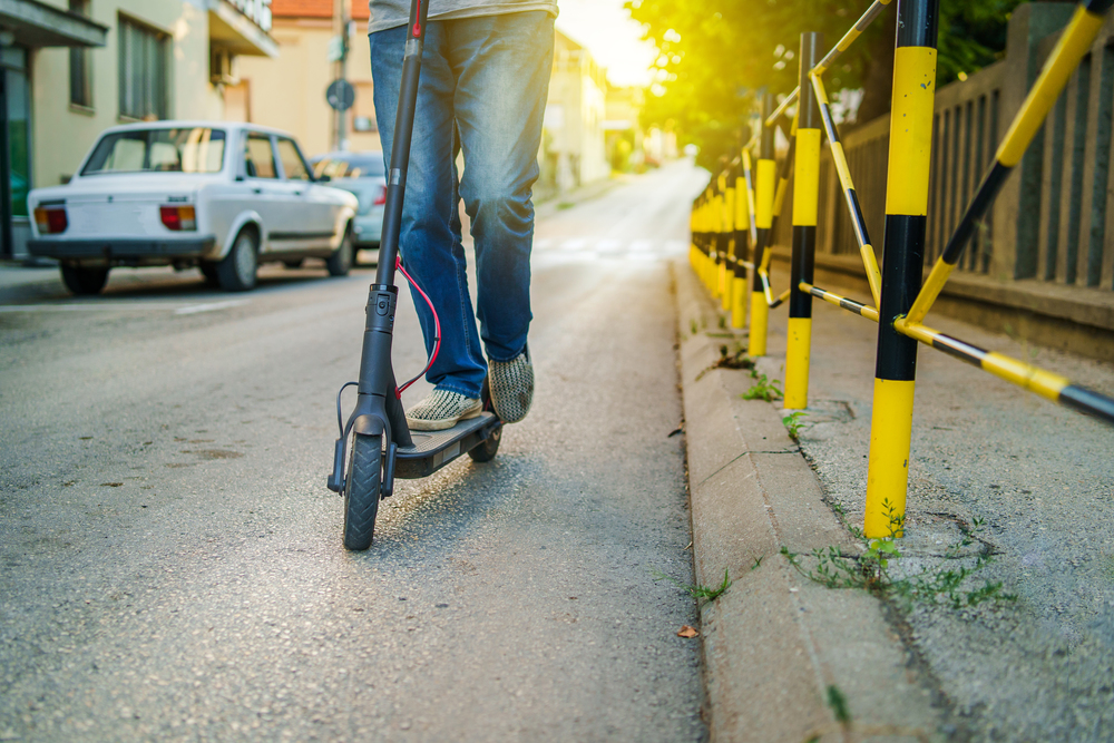 close up of senior man legs in jeans riding electric kick scooter on the street in summer evening day by the pavement fence