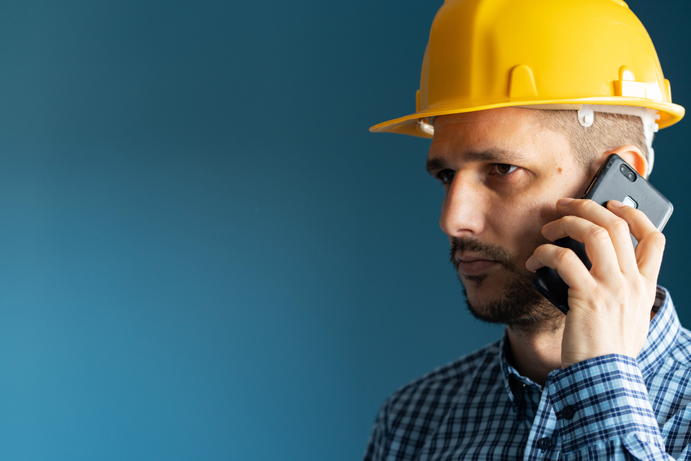 Close up portrait of young man engineer wearing yellow protecting safety helmet and shirt in front of blue wall isolated holding smart phone making a call talking side view