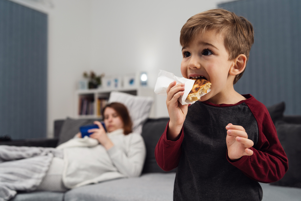 Portrait of small boy caucasian little child kid having dinner sandwich in napkin or pastry holding in front of opened mouth eating in the evening his mother is lying on bed in back plan defocused