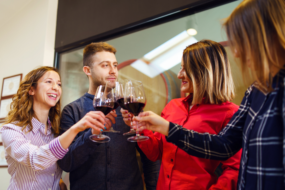 Group of caucasian friends or family standing by the table at winery or restaurant holding a glasses of red wine toasting celebrating young man and three women wearing shirt smiling