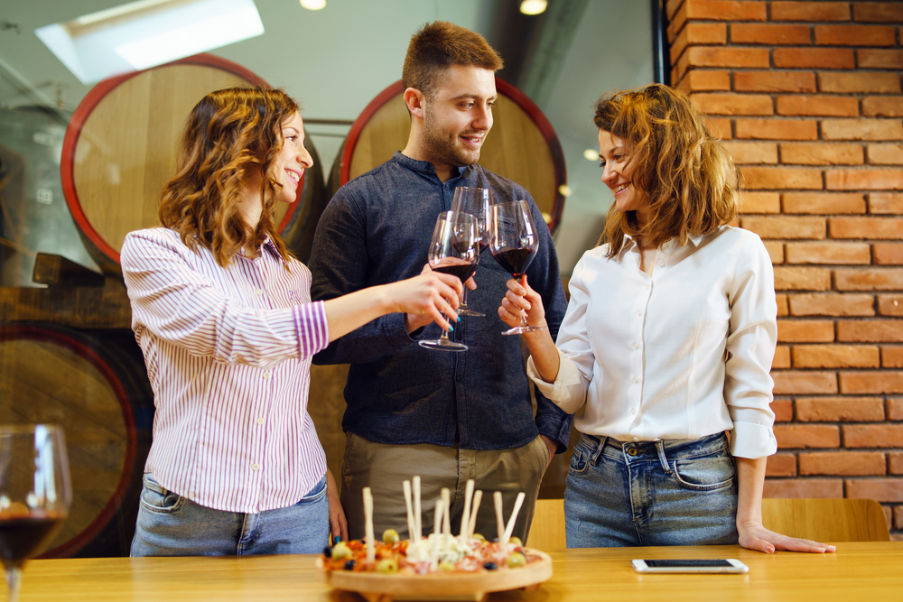 Caucasian friends two sisters or girlfriends and boyfriend with friend standing by the table at the restaurant or winery holding glasses of red wine toasting celebrating smiling