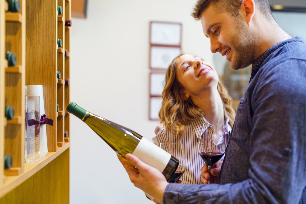 Young beautiful caucasian woman girl smiling in love while her boyfriend man is picking a wine bottle from the shelf reading holding glasses of red wine couple at winery or home husband and wife