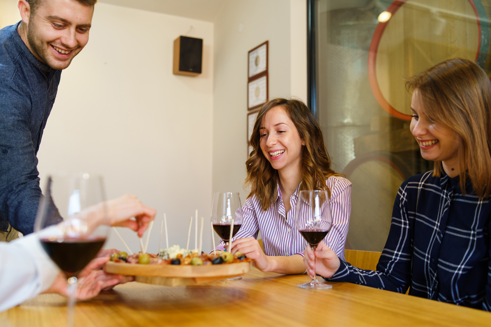 Two women caucasian girls sitting by the table holding glasses of red wine while one young man is serving appetizer friends smiling in day at home or restaurant