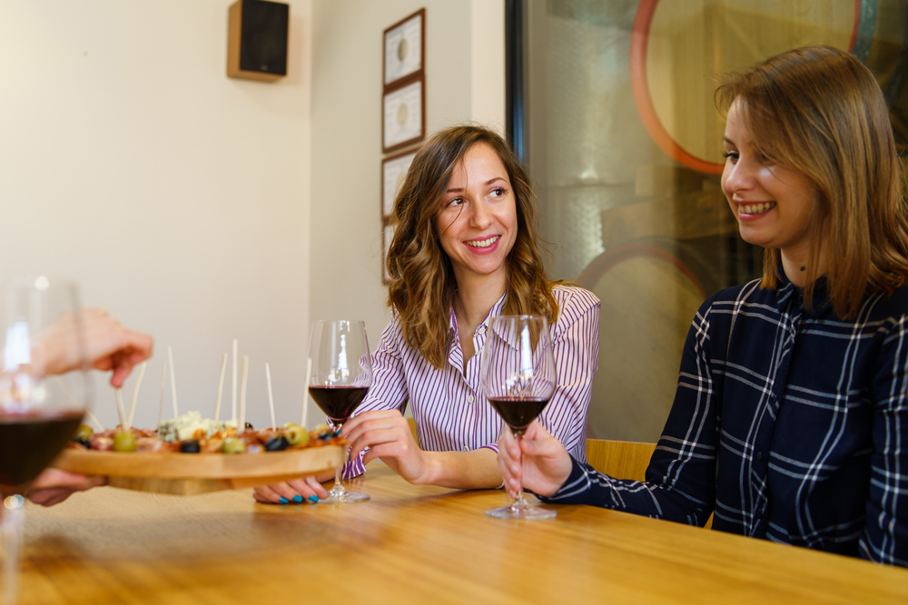 Two women caucasian girls sitting by the table holding glasses of red wine while unknown man is serving appetizer friends smiling in day at home or restaurant
