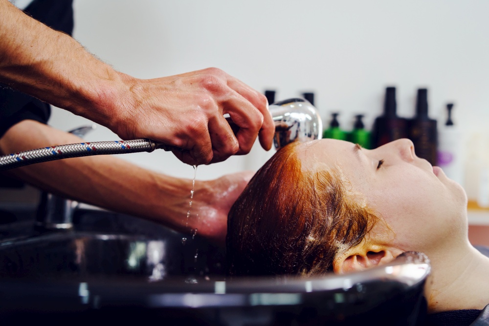 Close up on hand of adult caucasian unknown man male hairdresser holding shower head at the hairdressing salon while washing hair of unknown woman female black tub side view