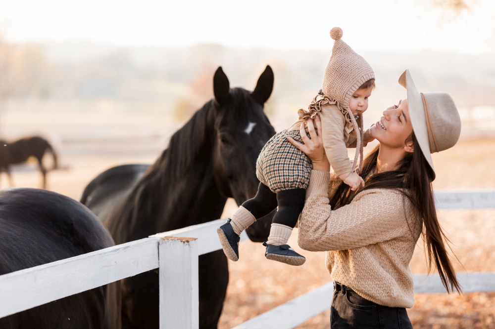 Young mother and little baby girl near a horses in autumn sunny day. Mother stroking a horse and smiling. mother&rsquo;s day.. Young mother and little baby girl near a horses in autumn sunny day. Mother stroking a horse and smiling. mother&rsquo;s day