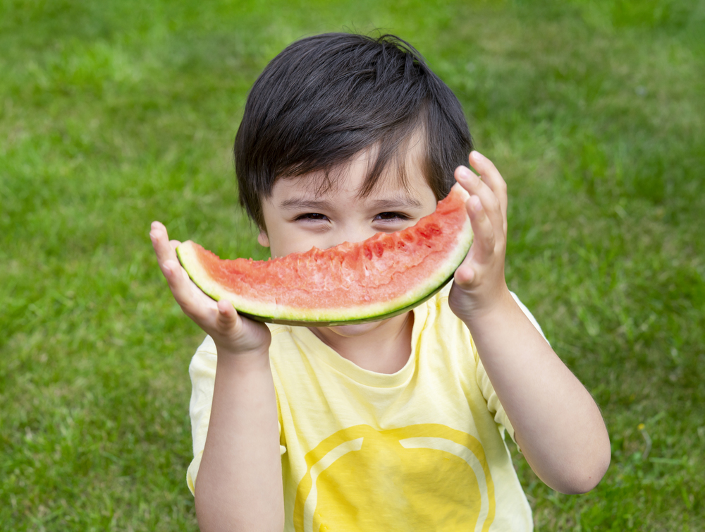 Selective focus Happy kid eating fresh watermelon outdoor in sunny day on summer, Hungry boy standing on grass eating watermelon looking at camera with smiling face, Healthy summer food for Children