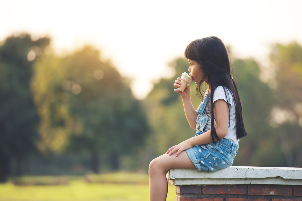 little children girl black hair white cloth sitting eating with ice cream on grren park background