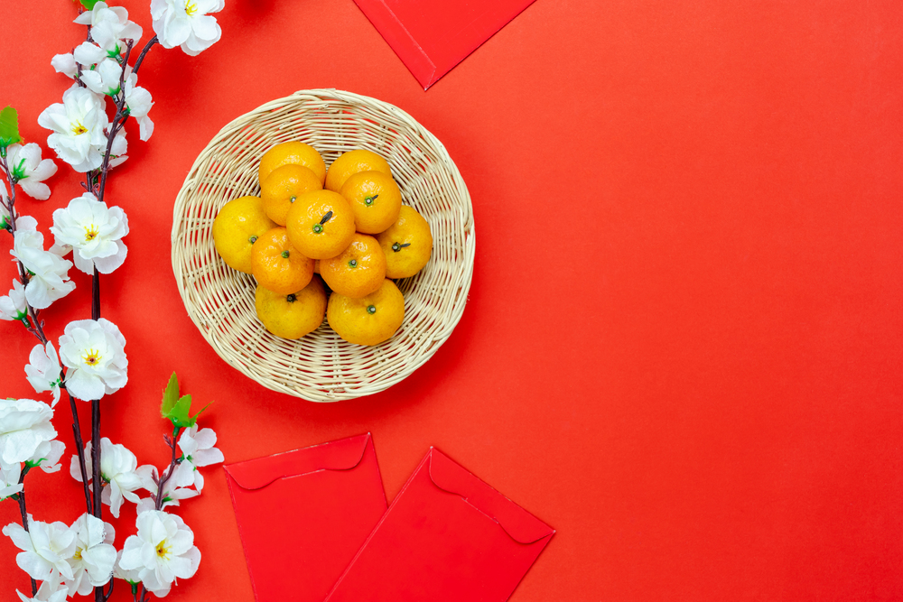 Table top view of accessories on Lunar New Year & Chinese New Year vacation concept background.Fresh orange in wood basket with white flower on modern rustic red backdrop at home office desk studio.