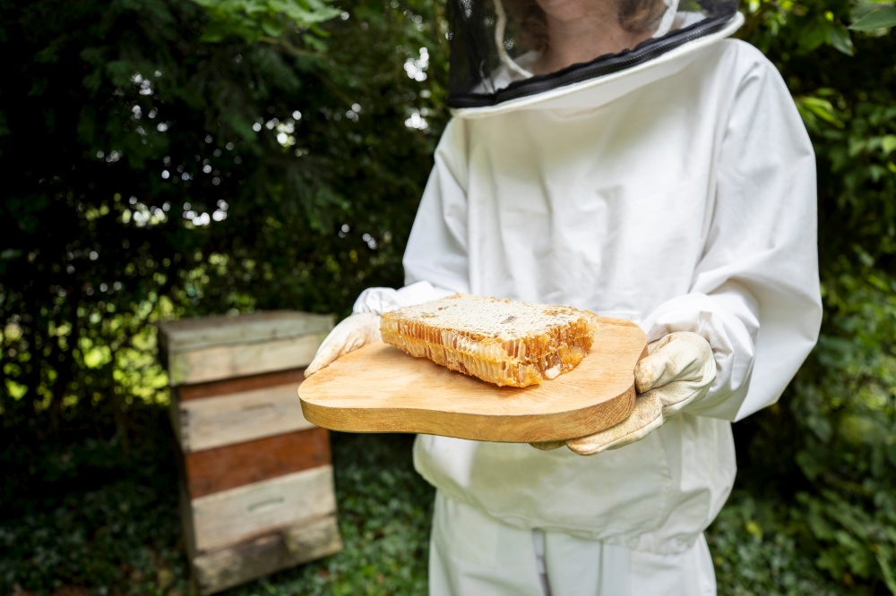 Female Beekeper In Protective Clothing Outside Honey Bee Hive Holding Honeycomb