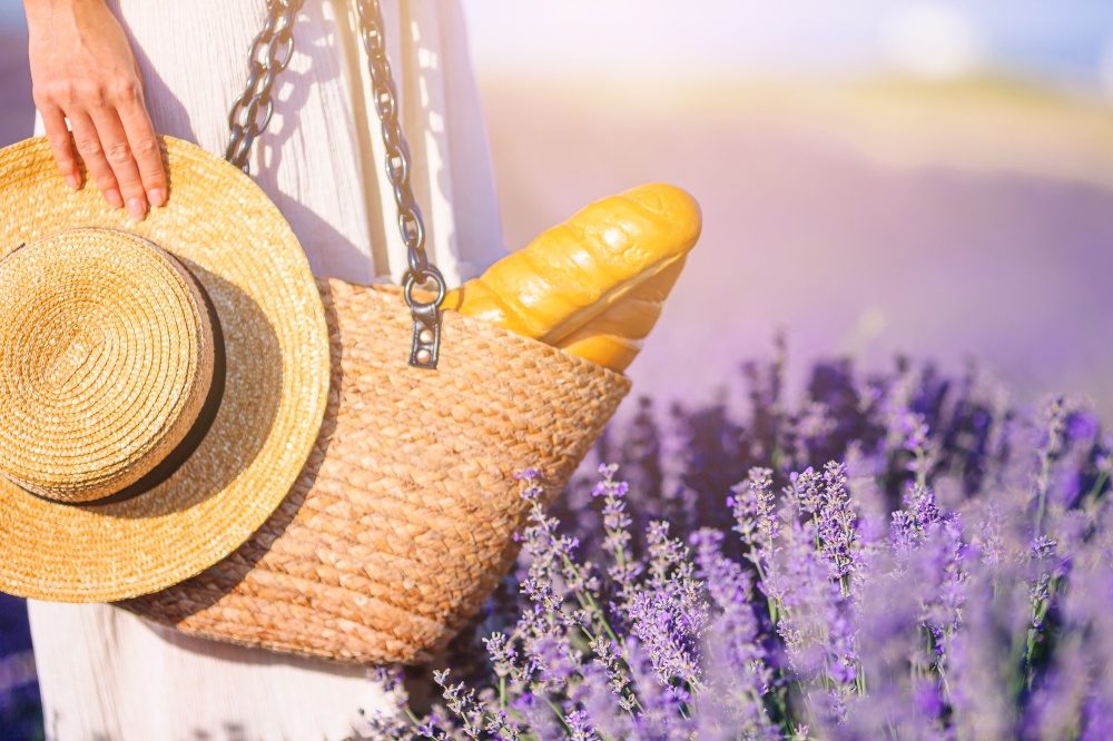 Closeup straw bag with bread and straw hat in female hands in lavender field. Closeup straw bag and hat in lavender field