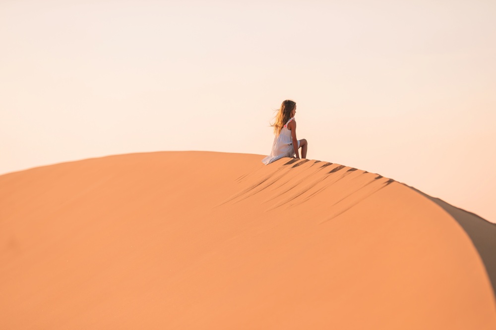 Little girl in white dress on vacation in big dunes in beautiful desert. Girl among dunes in desert in United Arab Emirates