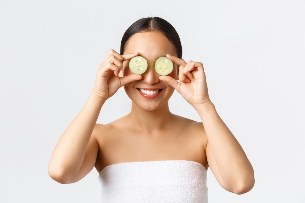 Beauty, personal care, spa salon and skincare concept. Close-up of beautiful smiling asian female in bath towel holding cucumber on eyes, promoting moisturzing, hydration cream or skin product.. Beauty, personal care, spa salon and skincare concept. Close-up of beautiful smiling asian female in bath towel holding cucumber on eyes, promoting moisturzing, hydration cream or skin product