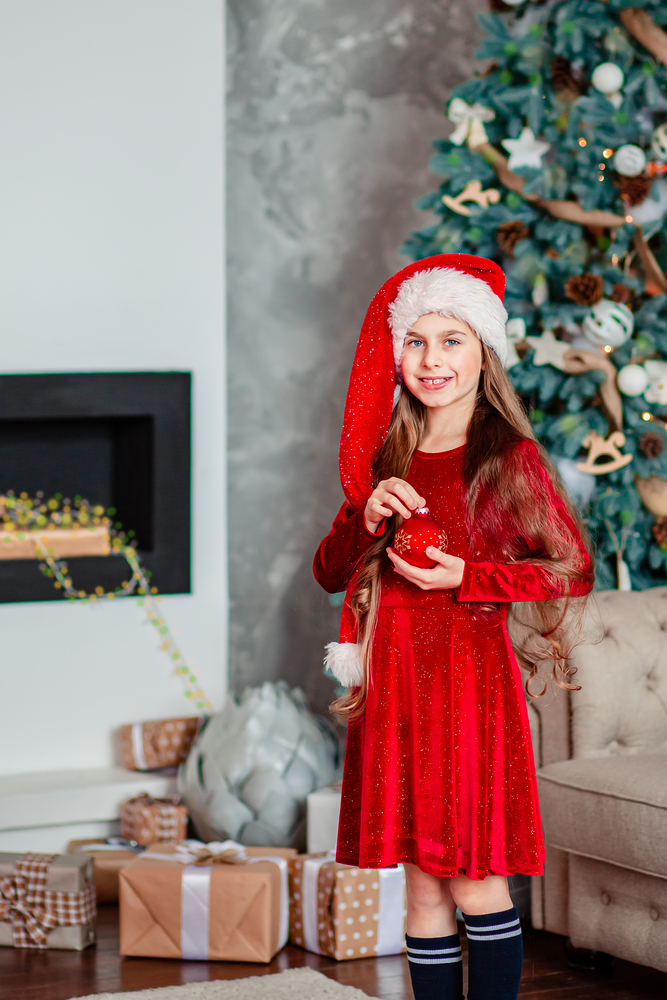One cute cheerful girl santa hat near Christmas tree. Holds red christmas ball. Merry Christmas.. One cute cheerful girl santa hat near Christmas tree. Holds red christmas ball.