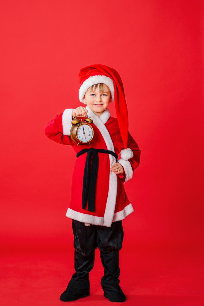 Little toddler boy dressed as Santa Claus holding a clock in his hand. Christmas shopping. A Christmas gift. Childhood. New Year party. Santa Claus baby. Happy winter holidays.. Little kid boy dressed as Santa Claus. Christmas shopping. Christmas present. Childhood. New Year party. Santa Claus baby.