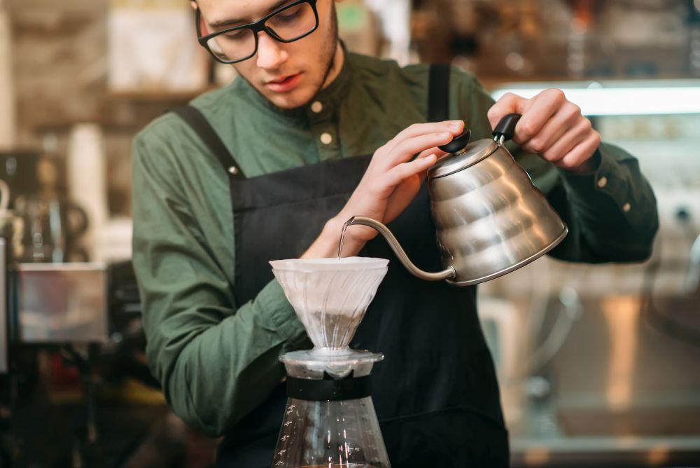 Waiter in black apron and eyeglasses pours hot boiled water in a coffee pot standing on bar counter.. Waiter pours hot boiled water in a coffee pot