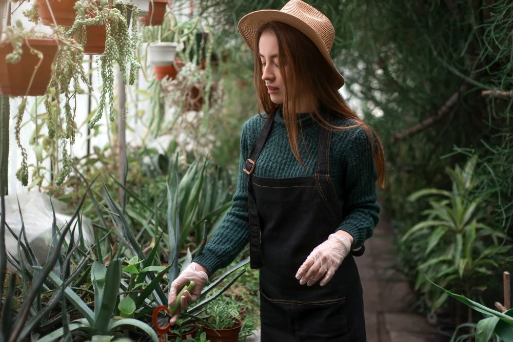 Young female florist in apron with shovel in hands against greenhouse on background. Female florist with shovel in hands