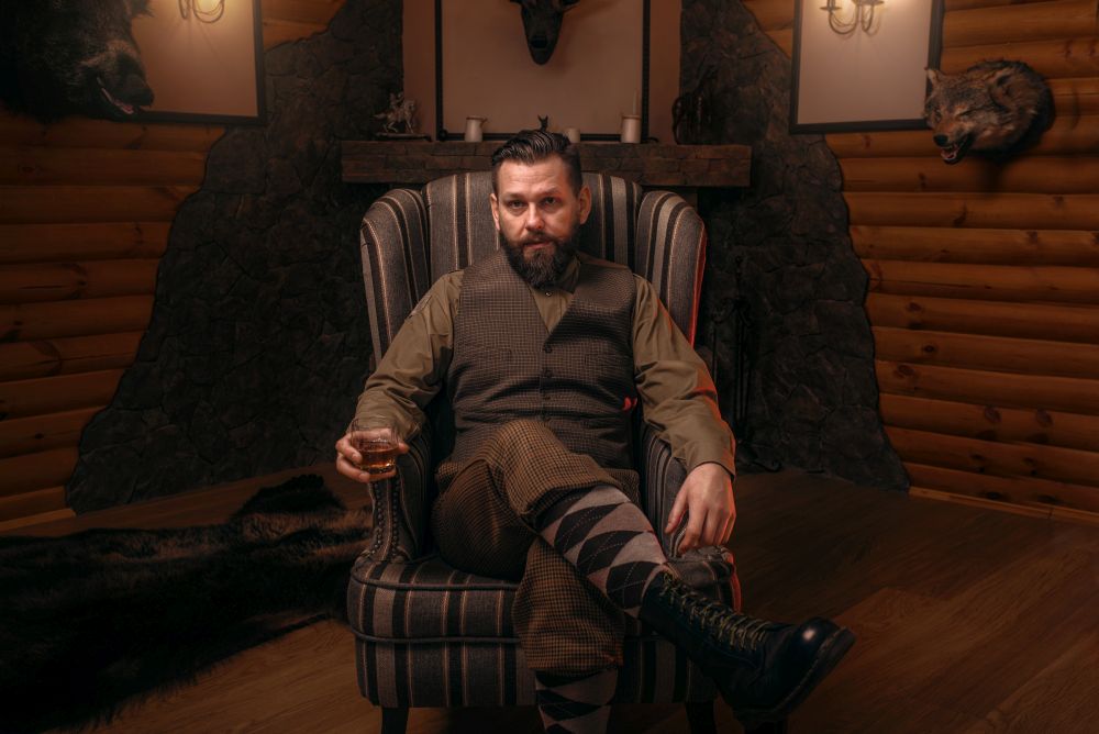 Hunter man in traditional british clothing sitting in a chair after hunting and drink whiskey. Fireplace, stuffed wild animals, bear skin and other trophies on background. Hunt lifestyle. Hunter man sitting in a chair and drink whiskey