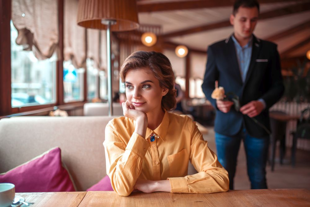 Beautiful woman waiting in restaurant, man with rose in hand on background