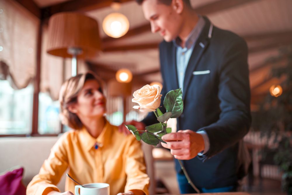 Man in suit gives rose flower to young happy woman, romantic date in restaurant. Beautiful relationship of love couple