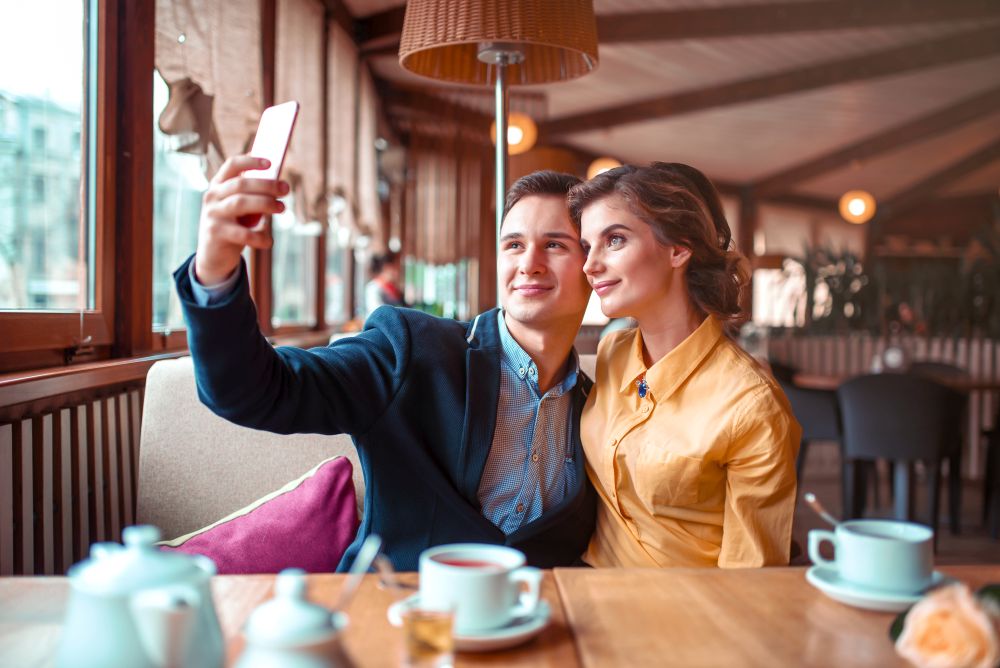 Love couple hugs and makes selfie on phone camera in restaurant. Man and woman happy together. Love couple hugs and makes selfie on phone camera
